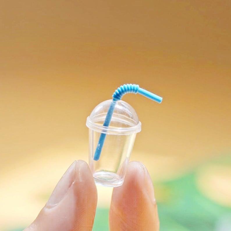 Puede incluir: Un vaso de pl&aacute;stico transparente en miniatura con una pajita azul. El vaso tiene una tapa y se sostiene entre dos dedos.