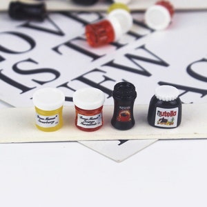May include: Four miniature glass jars with white lids. The jars contain orange marmalade, strawberry jam, Nutella spread, and Nescafe coffee. The jars are on a white surface with black letters.