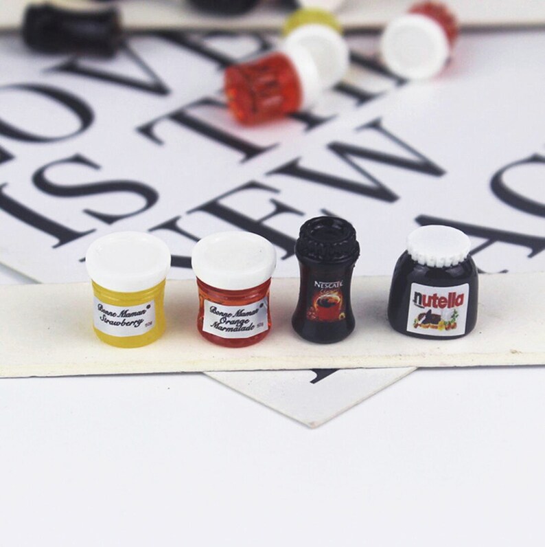 May include: Four miniature glass jars with white lids. The jars contain orange marmalade, strawberry jam, Nutella spread, and Nescafe coffee. The jars are on a white surface with black letters.