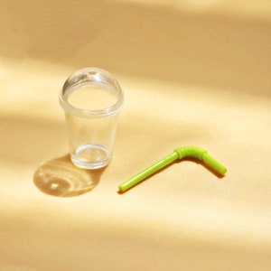 Puede incluir: Un vaso de pl&aacute;stico transparente con tapa y una pajita de pl&aacute;stico verde. El vaso y la pajita est&aacute;n sobre una superficie marr&oacute;n claro.