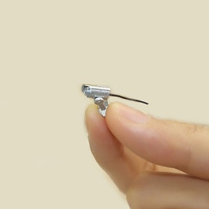 K&ouml;nnte beinhalten: Eine Miniatur-Sicherheitskamera aus Silber mit einem schwarzen Kabel. Die Kamera ist auf einem kleinen St&auml;nder montiert.