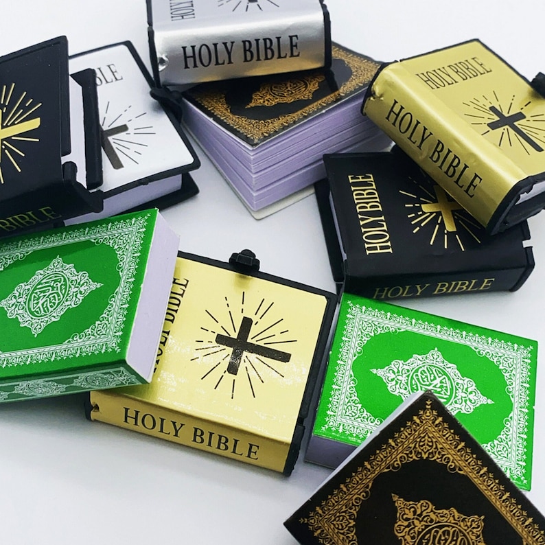 Puede incluir: Una colecci&oacute;n de Biblias en miniatura de varios colores y dise&ntilde;os. Las Biblias est&aacute;n hechas de papel y tienen una cubierta dorada o negra con un dise&ntilde;o de cruz. Las Biblias est&aacute;n dispuestas en una pila sobre una superficie blanca.