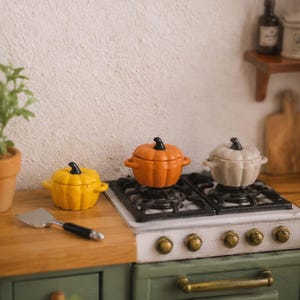 Könnte beinhalten: Miniatur-Küchenszene mit drei kürbisförmigen Töpfen in Gelb, Orange und Creme, jeweils mit schwarzem Griff. Die Töpfe stehen auf einem Miniaturherd, mit einem kleinen Spatel daneben. Die Szene hat ein Herbst- oder Erntethema.