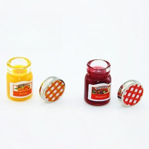 May include: Two miniature glass jars with lids, one filled with orange jam and labeled "Orange" and the other filled with red jam and labeled "Strawberry".