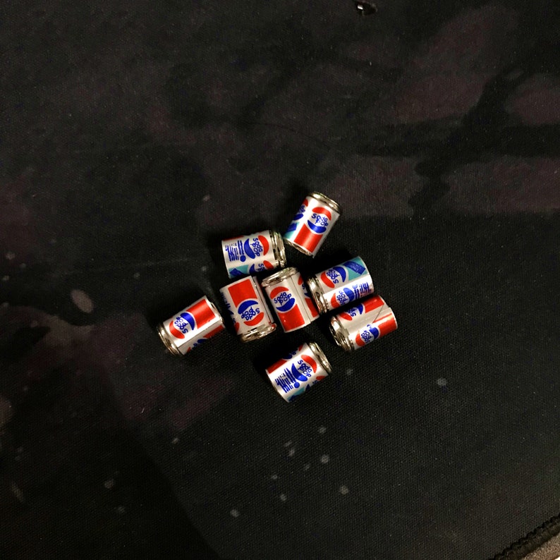May include: A group of eight small, silver, soda can-shaped charms with red, white, and blue designs. The charms are arranged in a loose circle on a black surface.