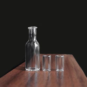 May include: A clear glass bottle with a narrow neck and two small, cylindrical glasses. The bottle and glasses are arranged on a wooden surface against a dark background. The glassware is transparent.