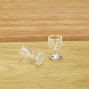 May include: Two miniature clear plastic wine glasses, one upright and one lying on its side, sit on a light brown wooden surface. The glasses have a classic stemmed design, perfect for a dollhouse or miniature collection.