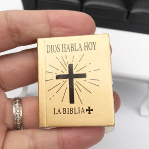 Puede incluir: Un peque&ntilde;o libro de color dorado con el texto "DIOS HABLA HOY LA BIBLIA" y una cruz negra con un dise&ntilde;o de sol.