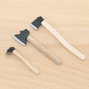 May include: Three miniature axes with wooden handles and dark grey axe heads. The axes vary in size, with the largest measuring approximately 15 cm in length. The axe heads have a silver-coloured edge.