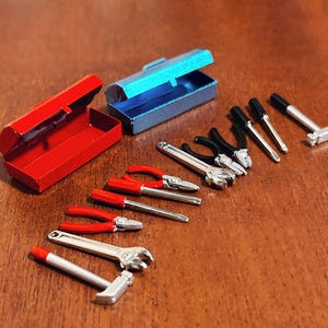 Può includere: Set di mini-utensili con una cassetta degli attrezzi rossa, una cassetta degli attrezzi blu e vari utensili come pinze, cacciaviti, una chiave inglese e un martello. Gli utensili hanno componenti in metallo argentato con dettagli neri e rossi.