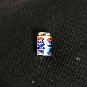 May include: A miniature silver can with a white label that says "Soda Pop" in blue letters. The can is on a black surface.