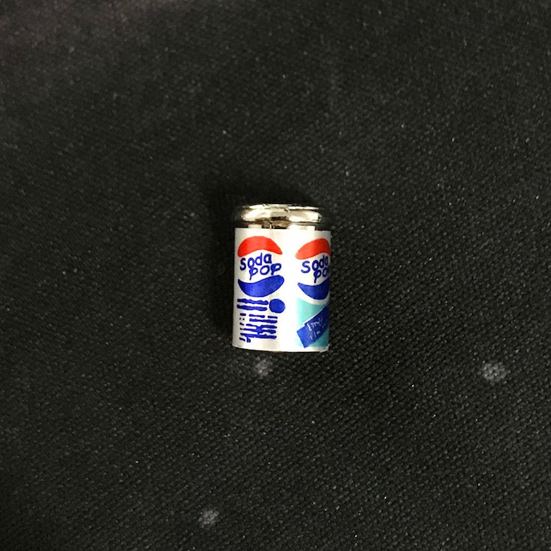 May include: A miniature silver can with a white label that says "Soda Pop" in blue letters. The can is on a black surface.