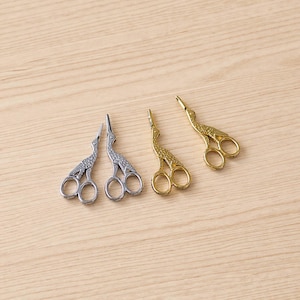 May include: Three pairs of decorative stork scissors in silver and gold tones. The scissors are arranged on a light wood surface. The scissors have a bird-like design, with the blades forming the beak and the handles forming the legs.