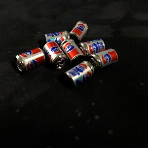 May include: A group of eight miniature silver cans with red and blue labels. The cans are arranged in a cluster on a black surface. The labels on the cans feature the words "Pepsi" and "Cola".