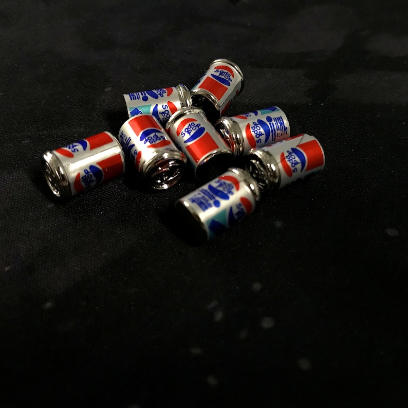 May include: A group of eight miniature silver cans with red and blue labels. The cans are arranged in a cluster on a black surface. The labels on the cans feature the words "Pepsi" and "Cola".