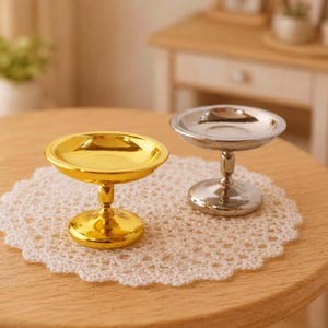 Puede incluir: Dos cuencos de pedestal en miniatura, uno dorado y otro plateado, se encuentran sobre un mantel de encaje blanco sobre una mesa de madera. El cuenco dorado tiene un acabado brillante, mientras que el cuenco plateado tiene un aspecto mate. Los cuencos son probablemente para fines decorativos.