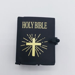 Puede incluir: Un llavero en forma de libro de imitaci&oacute;n de cuero negro con una cruz dorada y las palabras "HOLY BIBLE" impresas en la parte delantera.