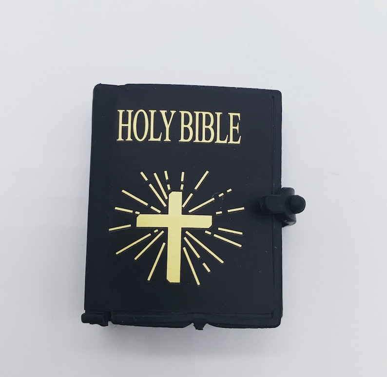 Puede incluir: Un llavero en forma de libro de imitaci&oacute;n de cuero negro con una cruz dorada y las palabras "HOLY BIBLE" impresas en la parte delantera.
