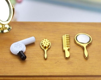 Op de afbeelding: Een set miniatuur gouden haarverzorgingstools voor een poppenhuis, inclusief een haarborstel, kam, handspiegel en föhn.