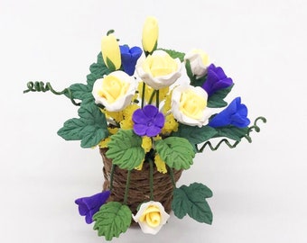 Puede incluir: Una pequeña cesta tejida de color marrón llena de un ramo de rosas amarillas, flores moradas y flores azules. Las flores están dispuestas de forma decorativa, con las rosas amarillas en el centro y las otras flores alrededor.