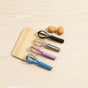 Può includere: Quattro piccole fruste in nero, argento, viola e blu, con manici a coste, sono disposte su una superficie di legno chiaro. Sono presenti anche due uova marroni e un piccolo tagliere in legno. Le fruste sono utensili da cucina.