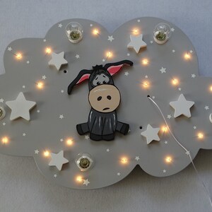 Op de afbeelding: Een grijs houten wolkvormig nachtlampje met een cartoon-ezel en witte sterren. Het lampje heeft een string met kleine witte lampjes rond de rand.