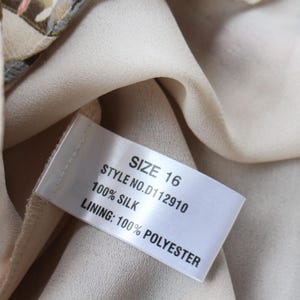 May include: Close-up of a garment tag with the text "SIZE 16", "STYLE NO. D112910", "100% SILK", and "LINING: 100% POLYESTER". The tag is attached to a cream-colored fabric. The garment has a patterned design with red and beige tones.