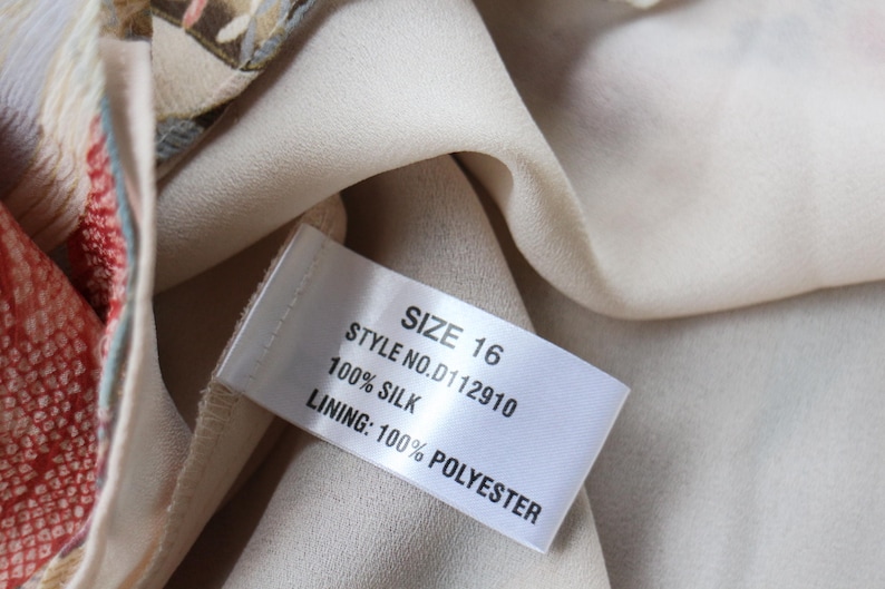 May include: Close-up of a garment tag with the text "SIZE 16", "STYLE NO. D112910", "100% SILK", and "LINING: 100% POLYESTER". The tag is attached to a cream-colored fabric. The garment has a patterned design with red and beige tones.