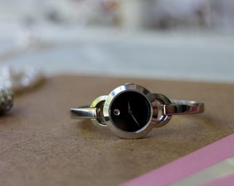 Puede incluir: Un reloj de pulsera plateado con esfera negra y un pequeño detalle rosa. El reloj tiene un diseño minimalista con una correa de estilo eslabones. El reloj se muestra sobre una superficie marrón claro.
