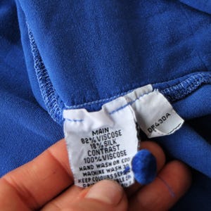 May include: Close-up of a vibrant blue fabric with a sewn seam. A white tag is attached, displaying text including "MAIN 82% VISCOSE 18% SILK" and "CONTRAST 100% VISCOSE". The tag also includes washing instructions and a product code.