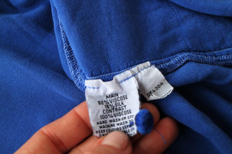 May include: Close-up of a vibrant blue fabric with a sewn seam. A white tag is attached, displaying text including "MAIN 82% VISCOSE 18% SILK" and "CONTRAST 100% VISCOSE". The tag also includes washing instructions and a product code.