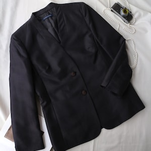 May include: A black blazer with a mandarin collar and two-button closure. The jacket is displayed on a white surface, with a pearl necklace and a vintage camera nearby. The blazer has a tailored fit and is made of a smooth fabric.