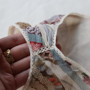 May include: Close-up of a cream-colored garment with a patterned neckline. The neckline features a patchwork design with blue, red, and floral patterns, trimmed with delicate lace. The garment is held by a hand with a gold ring.