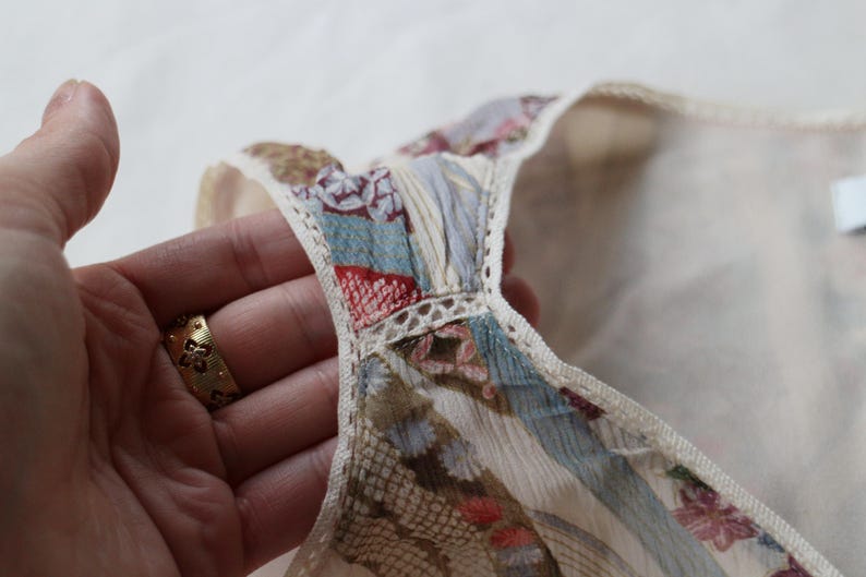 May include: Close-up of a cream-colored garment with a patterned neckline. The neckline features a patchwork design with blue, red, and floral patterns, trimmed with delicate lace. The garment is held by a hand with a gold ring.