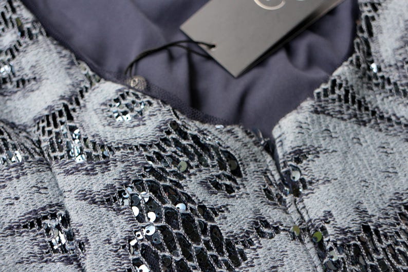 May include: Close-up of a gray and black sequined top. The fabric has a textured pattern with silver sequins. A gray tag hangs from the garment. The top is likely part of a formal wear collection.