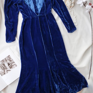 May include: A long, royal blue velvet dress with long sleeves and a plunging neckline. The dress is displayed on a white surface, with a pearl necklace and a white flower with red accents. A book is open in the lower left corner.