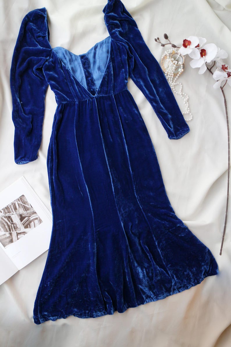 May include: A long, royal blue velvet dress with long sleeves and a plunging neckline. The dress is displayed on a white surface, with a pearl necklace and a white flower with red accents. A book is open in the lower left corner.