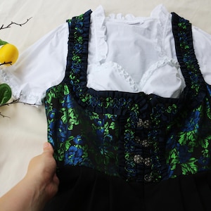 K&ouml;nnte beinhalten: Ein traditionelles deutsches Dirndl-Kleid. Das Oberteil ist marineblau mit einem gr&uuml;nen und blauen Blumenmuster, und das Kleid hat einen schwarzen Rock. Eine wei&szlig;e Bluse mit R&uuml;schen&auml;rmeln und Ausschnitt ist darunter zu sehen.