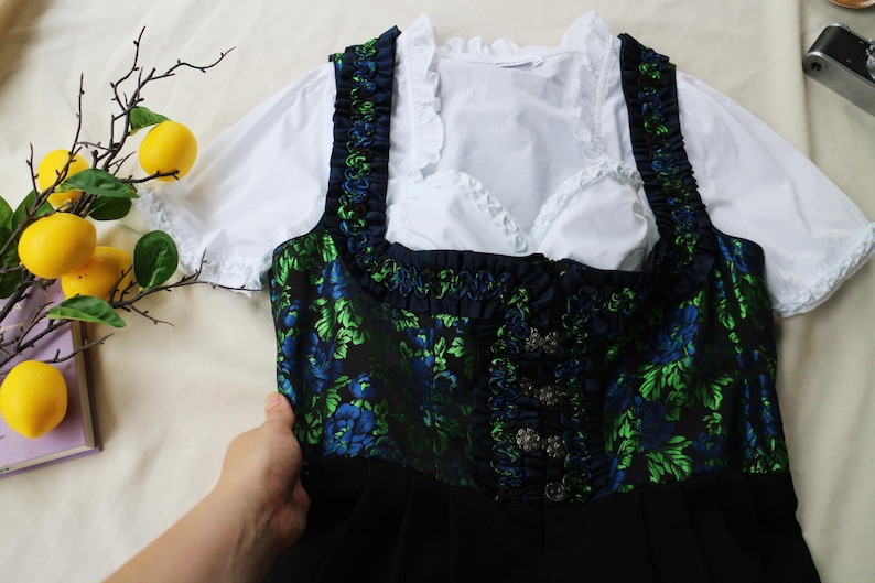 K&ouml;nnte beinhalten: Ein traditionelles deutsches Dirndl-Kleid. Das Oberteil ist marineblau mit einem gr&uuml;nen und blauen Blumenmuster, und das Kleid hat einen schwarzen Rock. Eine wei&szlig;e Bluse mit R&uuml;schen&auml;rmeln und Ausschnitt ist darunter zu sehen.