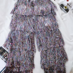 Könnte beinhalten: Eine Hose mit mehreren horizontalen Lagen aus irisierenden, metallischen Fransen in Silber-, Lila-, Blau- und Grüntönen. Das Kleidungsstück hat einen hohen Bund und eine geschichtete, zottelige Textur, die die gesamte Beinlänge bedeckt.