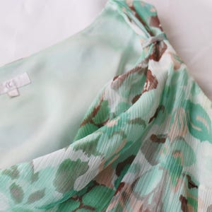 May include: A close-up of a light green and brown patterned dress. The dress has a flowing, sheer fabric with a subtle texture. The design includes a knot detail at the shoulder. The inner lining is a solid light green color.
