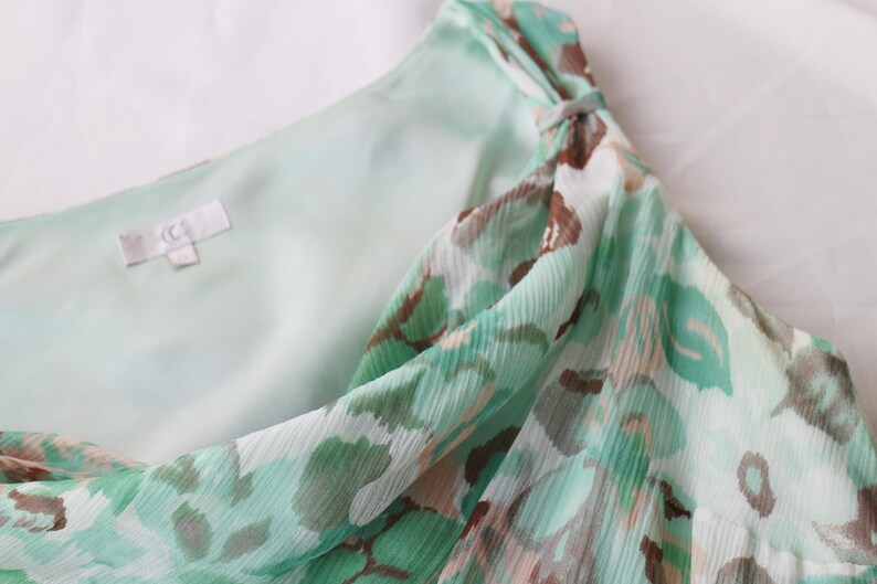 May include: A close-up of a light green and brown patterned dress. The dress has a flowing, sheer fabric with a subtle texture. The design includes a knot detail at the shoulder. The inner lining is a solid light green color.