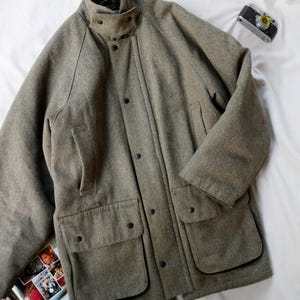 May include: A taupe-coloured wool-blend jacket featuring a high collar with a snap-button throat latch, a front placket with snap closures, two large flap pockets at the waist, and two vertical slit pockets on the chest with long sleeves.