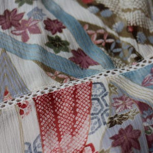 May include: Close-up of a patterned fabric with a mix of floral and geometric designs. The fabric features a palette of cream, red, blue, and brown hues. Delicate lace trim accents the seams, adding a touch of elegance to the textile.