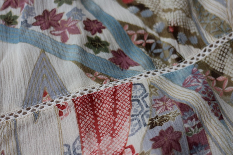 May include: Close-up of a patterned fabric with a mix of floral and geometric designs. The fabric features a palette of cream, red, blue, and brown hues. Delicate lace trim accents the seams, adding a touch of elegance to the textile.