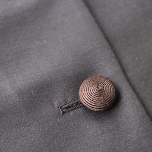 May include: Close-up of a dark gray fabric with a brown, spiral-patterned button and buttonhole. The button is attached to the fabric, which appears to be part of a garment, possibly a coat or jacket.