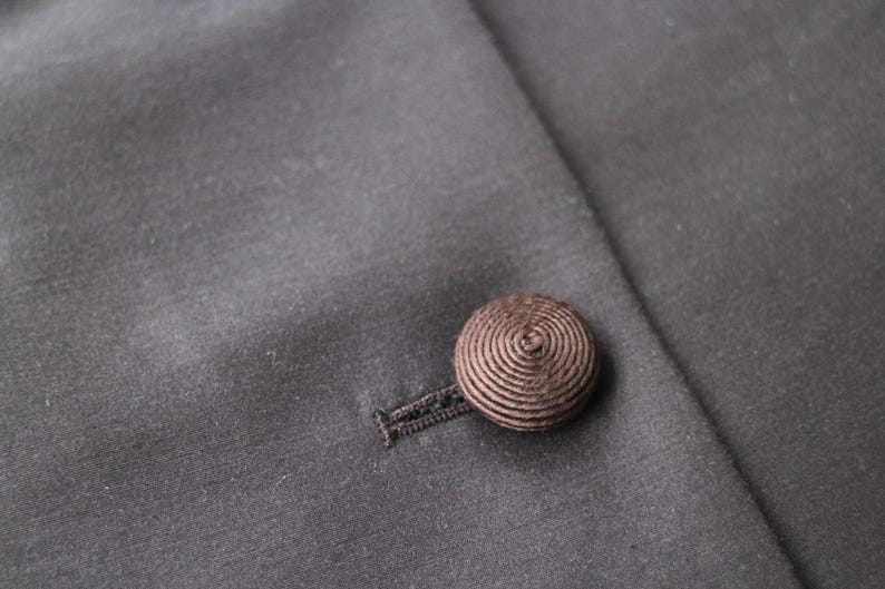 May include: Close-up of a dark gray fabric with a brown, spiral-patterned button and buttonhole. The button is attached to the fabric, which appears to be part of a garment, possibly a coat or jacket.