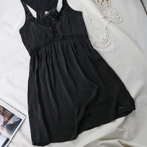 May include: A black sleeveless tank top featuring a racerback design and a scoop neckline. The garment is adorned with intricate black beadwork along the straps and neckline, with a pleated bodice that flows into a loose, flared hemline.