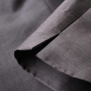 May include: Close-up of dark gray fabric with a visible seam. The fabric appears to be smooth and possibly woven, with subtle variations in tone. The seam creates a sharp, dark line.