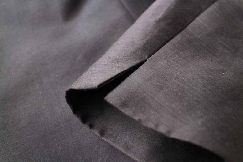 May include: Close-up of dark gray fabric with a visible seam. The fabric appears to be smooth and possibly woven, with subtle variations in tone. The seam creates a sharp, dark line.
